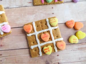 three graham crackers with pink or white frosting on it and candy conversation hearts to look like a tic tac toe board on a wood table with more candy next to it 