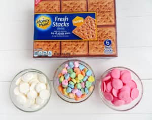 a box of graham crackers, bowls of white and pink candy melts and conversation heart candy on a white wood table