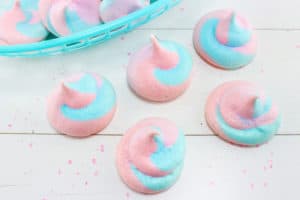 pink and blue meringues in a blue basket and in front of the basket on a white wood table