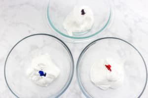 three glass bowls of meringue with a drop of food coloring in them on a counter