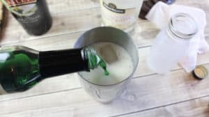 pouring alcohol into a metal glass with more bottles of alcohol in the background on a wood table