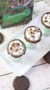 an overhead view of two thin mint shots in shot glasses rimmed with chocolate on a white plate on a wood table with a box of thin mints cookies in the background 