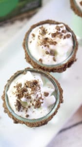 an overhead view of two thin mint shots in shot glasses rimmed with chocolate on a white plate on a wood table with a box of thin mints cookies in the background 