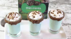 three thin mint shots in shot glasses rimmed with chocolate on a white plate on a wood table with a box of thin mints cookies in the background 
