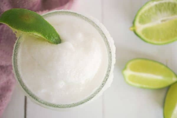 an overhead view of a coconut lime margarita in mason jar with lime wedge garnish and more lime wedges and a pink dishcloth in background on a white wood table