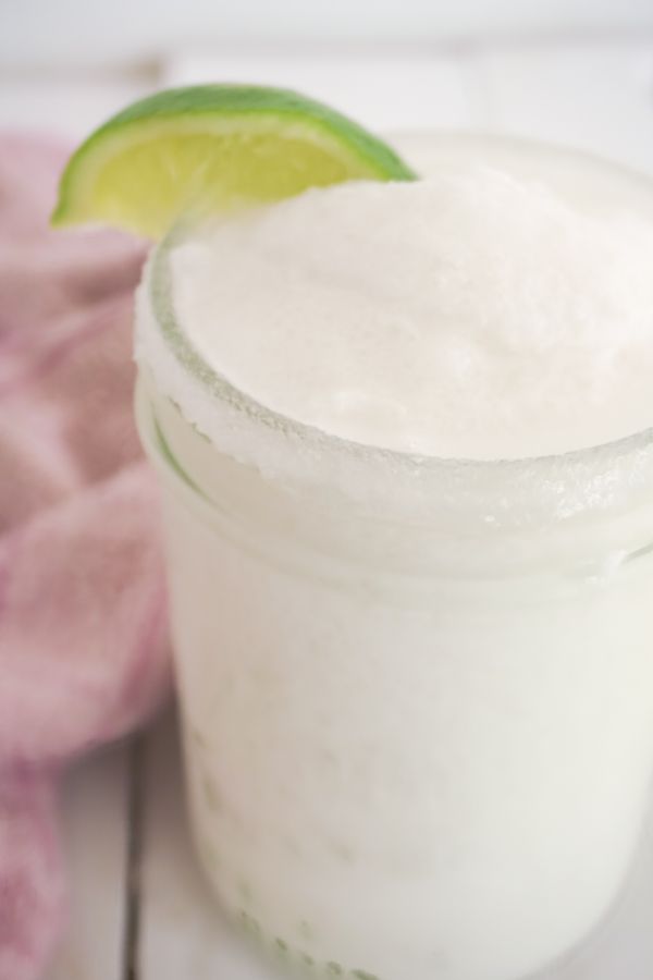 Close up of coconut lime margarita in mason jar with lime wedge garnish and pink dishcloth in background on a white wood table