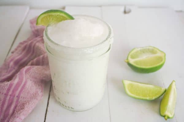 coconut lime margarita in mason jar with lime wedge garnish and more lime wedges and a pink dishcloth in background on a white wood table