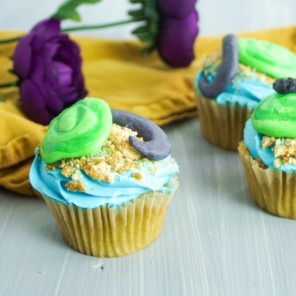 three moana cupcakes decorated to look like the Heart of Te Fiti with green, blue and gray frosting on a grey background with yellow linen and purple flower in the background