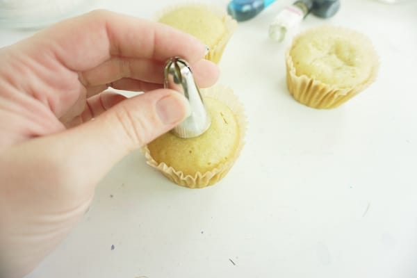hand pushing an icing tip into a cupcake to create a well with two more cupcakes in the background on a white table
