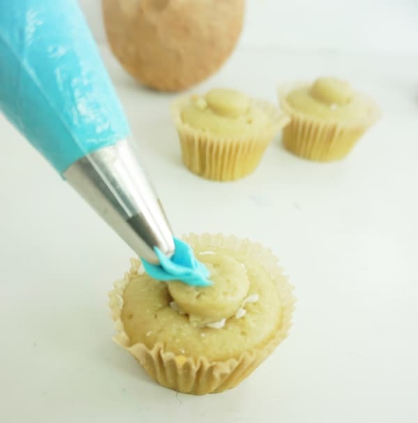 blue icing being placed on a cupcake from a pastry bag and decorating tip with more cupcakes in the background on a white table