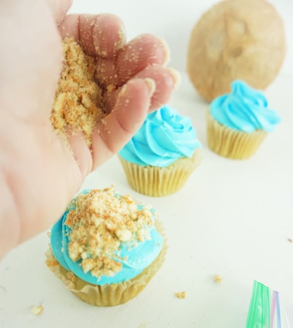 hand pouring crushed vanilla wafer cookie on a blue iced cupcake with more iced cupcakes in the background on a white table
