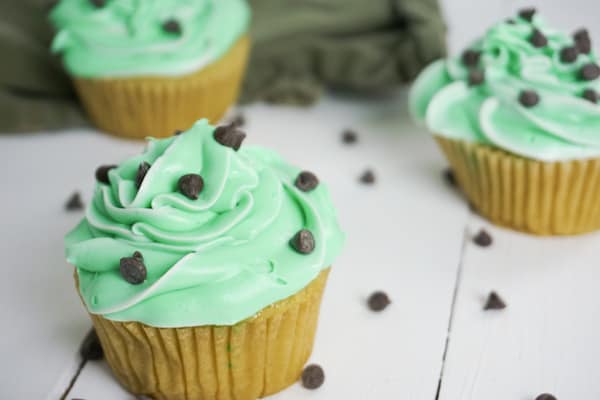 side view of three boozy mint cupcakes with chocolate chips on a white table and a green linen in the background