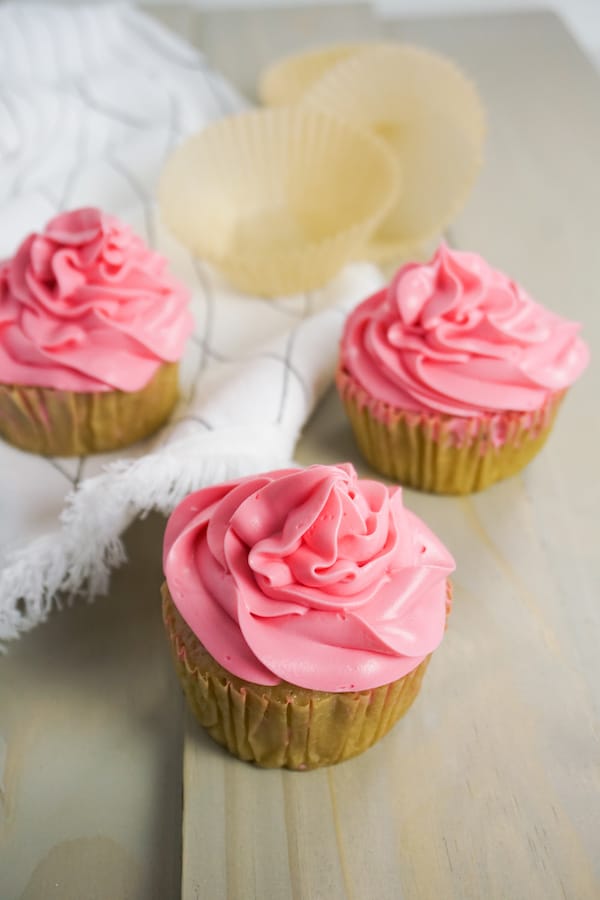overhead view of three strawberry boozy cupcakes on a grey wood table with a white linen and cupcake liners in the background