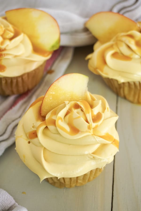 three cupcakes topped with caramel frosting, caramel sauce and an apple slice on a brown table with a brown linen in the background