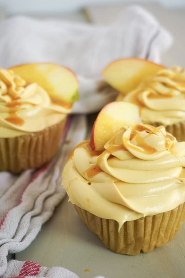 three cupcakes topped with caramel frosting, caramel sauce and an apple slice on a brown table with a brown linen in the background