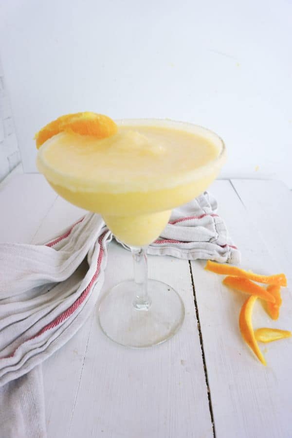 orange creamsicle margarita in a glass with an orange slice on the edge with orange rinds and a white and red cloth next to it on a white wood table
