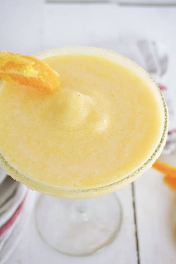a closeup view of an orange creamsicle margarita in a glass with an orange slice on the edge with orange rinds and a white and red cloth next to it on a white wood table
