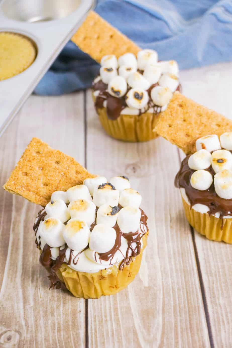 side view of three cupcakes frosted with chocolate frosting topped with marshmallows and a graham cracker on a wood background with a muffin tin and blue linen in the background