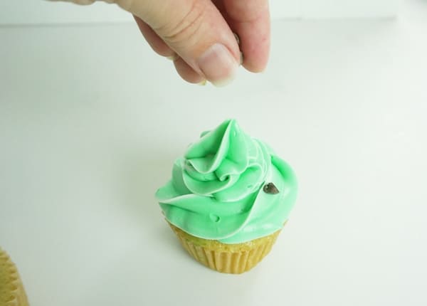hand dropping chocolate chips on top of green iced cupcake on a white table