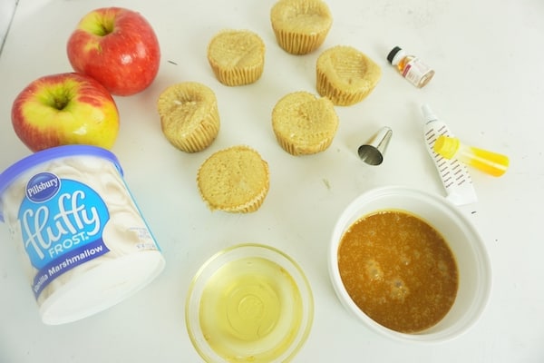 apples, white frosting, cupcakes, a bowl of caramel sauce, decorating tip, food coloring and gel, all on a white table