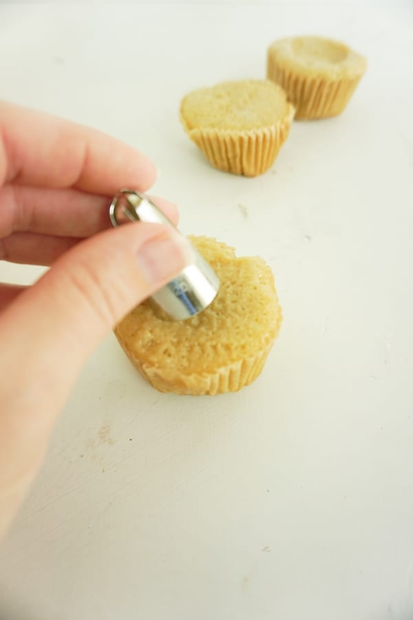 a hand placing a decorating tip in the middle of a cupcake on a white table with more cupcakes in the background