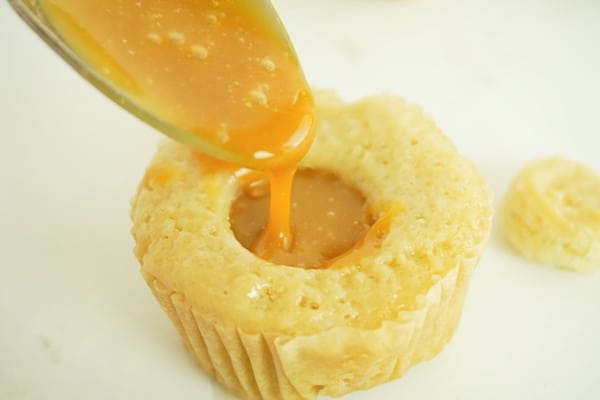 a spoon pouring caramel sauce into the middle of a cupcake on a white table