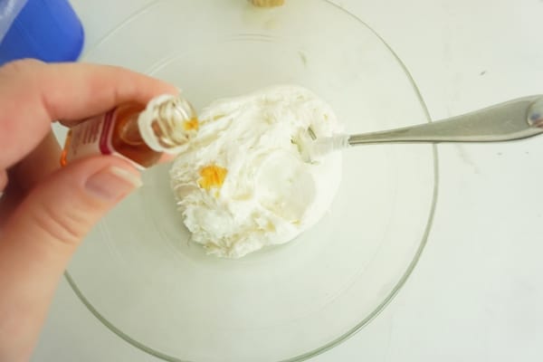 a hand dropping some caramel flavoring into a bowl of white frosting with a spoon in it, on a white table