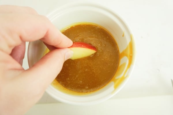 a hand dipping an apple slice in a bowl of caramel sauce on a white table