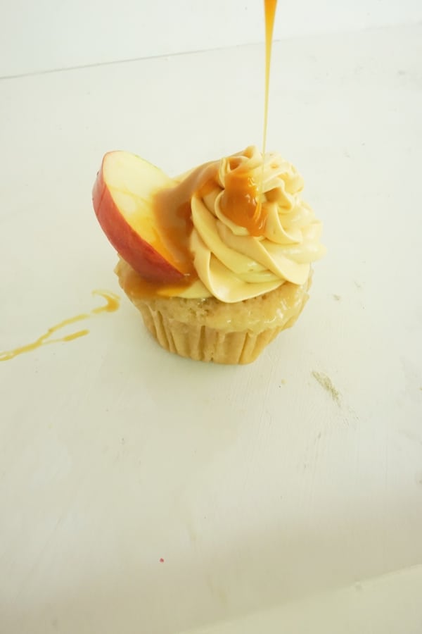 caramel sauce being drizzled from above on a cupcake with caramel frosting and an apple slice on a white table