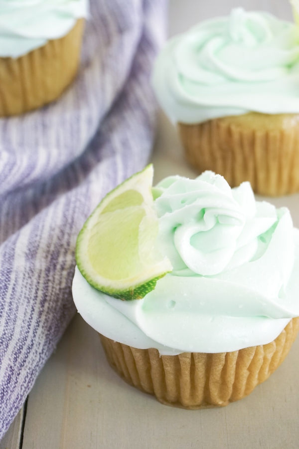 Tequila-Infused Margarita Cupcakes topped with green frosting and a slice of lime on a wood table next to a purple linen