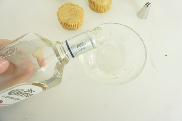 tequila being poured into a glass bowl with cupcakes and a decorating tip in the background on a white table