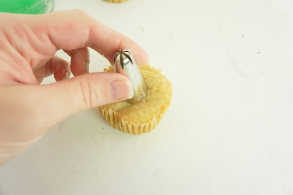 hand pushing a piping tip into the top of a cupcake to make a well inside the cupcake on a white table