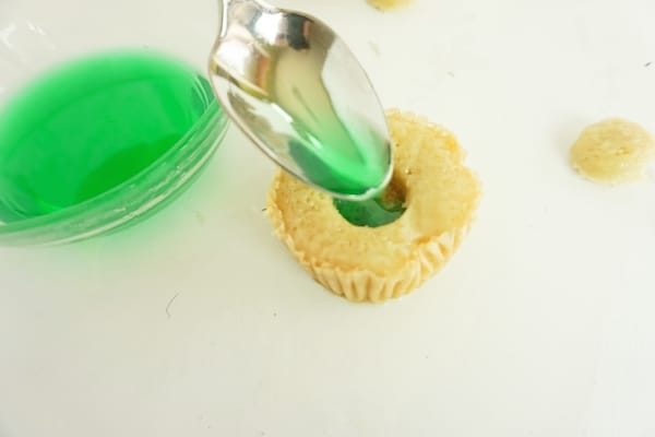 spoon filling a cupcake with green colored tequila mixture to make a margarita cupcake on a white table
