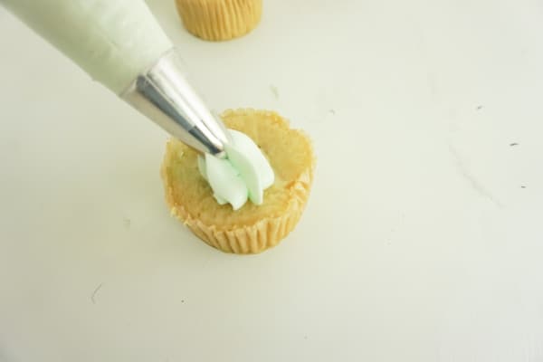 light green icing being placed onto a cupcake with a piping bag on a white table