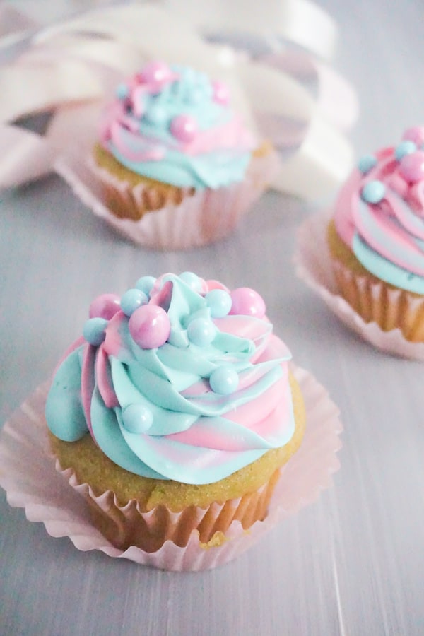 side view of three cupcakes frosted with pink and blue frosting with pink and blue large sugar pearls on a grey table with white pink and grey ribbons in the background