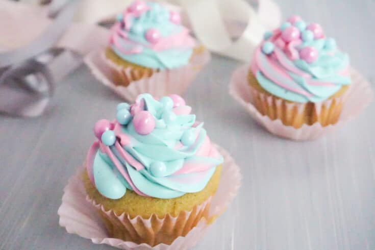 side view of three pink and blue swirled cupcakes with pink and blue large sugar pearls on a grey table with white pink and grey ribbons in the background