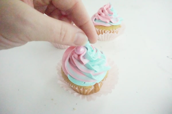hand placing large sugar pearls on pink and blue swirled cupcake with another cupcake in the background, all on a white table