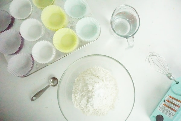 a muffin tin with cupcake liners in it, a measuring cup of water, a measuring spoon, a bowl of flour, a whisk  on a white table