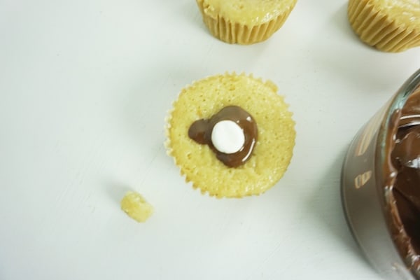 chocolate oozing out of the top of a cupcake around a mini-marshmallow stuffed into a well in the cupcake with more cupcakes and a bowl of melted chocolate on a white table in the background