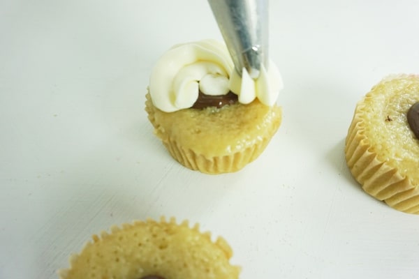 decorating tip placing white icing on top of a chocolate-filled cupcake with two more cupcakes in the background on a white table