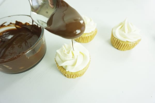 spoon drizzling chocolate over the top of a cupcake with white icing with two more cupcakes with white icing and a bowl of melted chocolate in the background on a white table