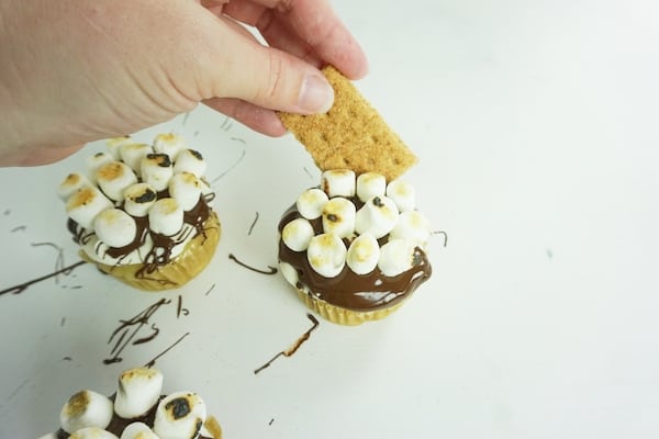 hand placing a piece of graham cracker on top of a cupcake topped with melted chocolate and marshmallows next to two more cupcakes on a white table