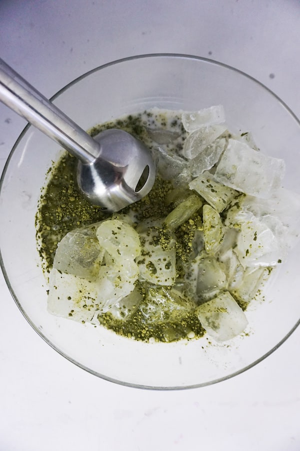 ice and matcha milk mixture being blended with an immersion blender in a bowl on a white table