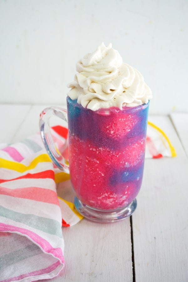 bright pink and blue unicorn frappuccino recipe in a glass mug with whipped cream on top next to brightly stripped linen on a white background