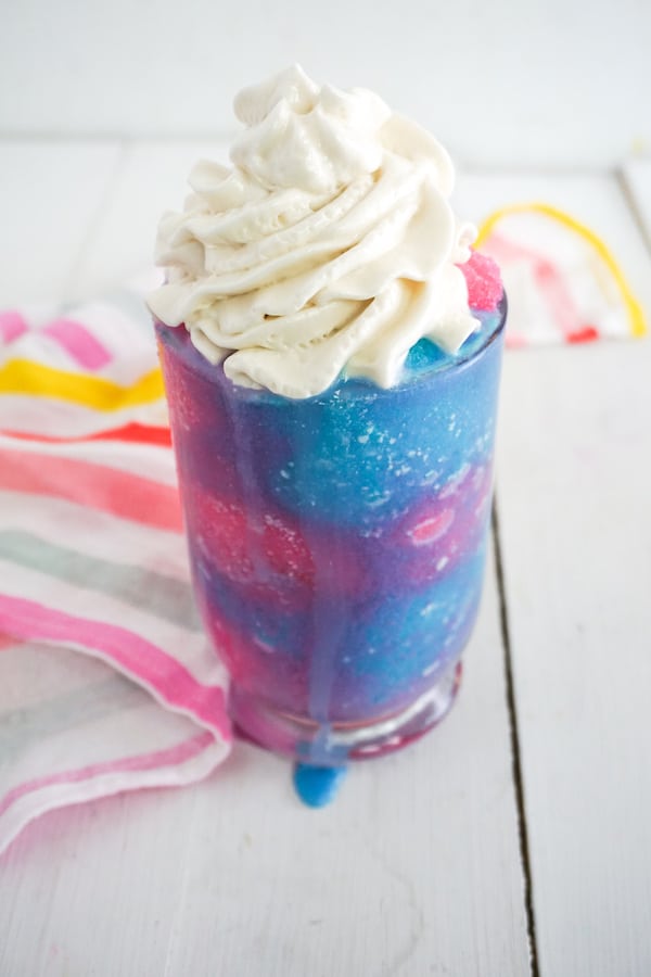 brightly colored pink and blue chocolate flavored frappuccino topped with whipped cream on a white background with a rainbow striped linen