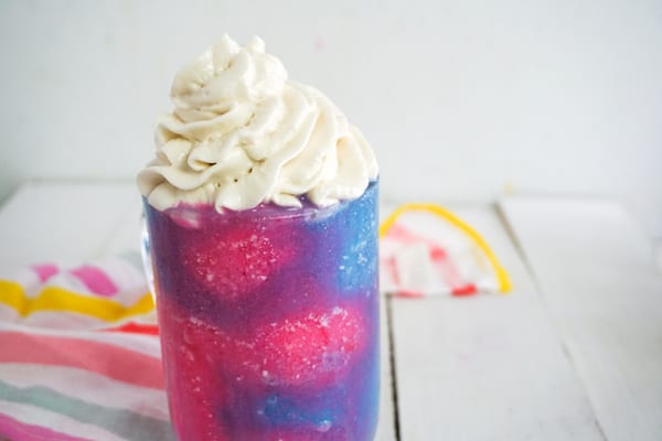 side view of pink and blue unicorn frappuccino topped with whipped cream on a white background with a brightly striped linen