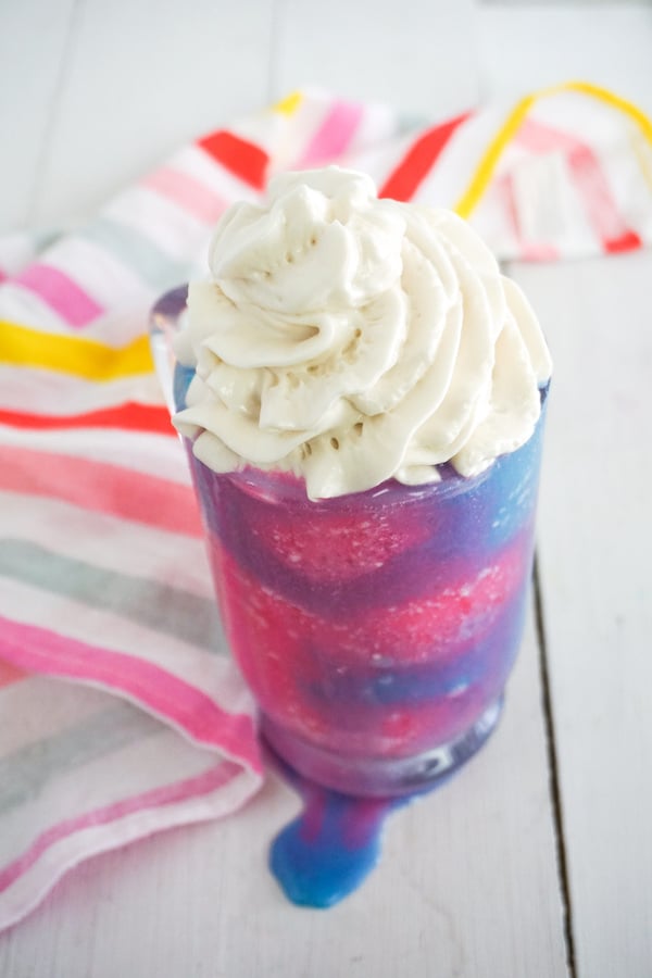 bright blue and pink frappuccino with whipped cream on top in a glass mug on a white table with brightly striped linen