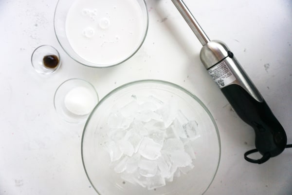 glass bowls of milk, vanilla bean paste, sugar and ice on a white table next to an immersion blender