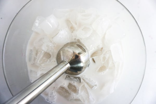 ice and milk being blended in a glass bowl with an immersion blender on a white table to make a vanilla bean frappuccino