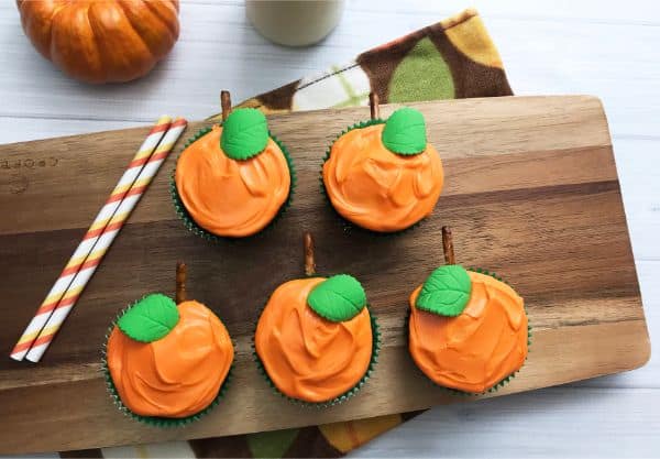 chocolate pumpkin cupcakes decorated with orange frosting and a green candy leaf, all on a wooden cutting board on a white wood table with a pumpkin in the background